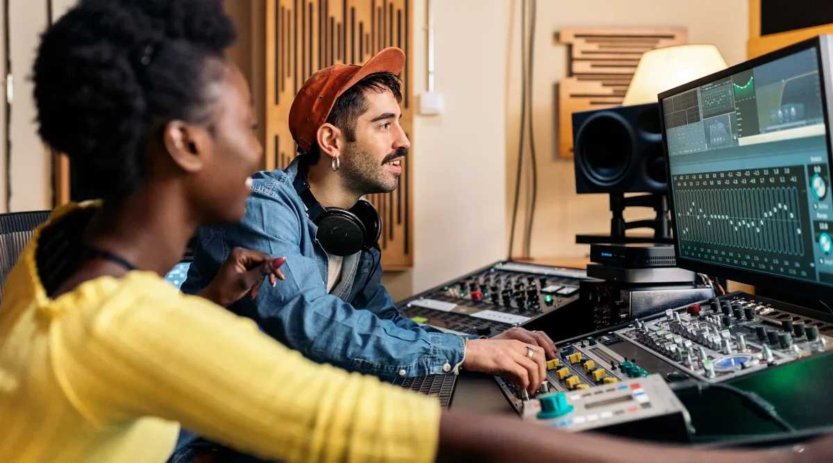 A studio engineer sits at a soundboard and listens to playback on a recording while a woman looks on.