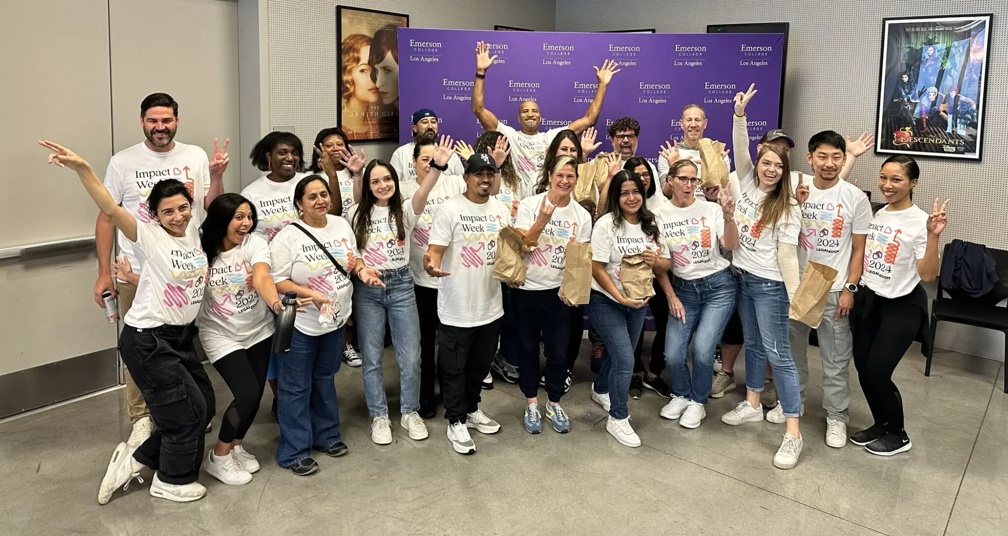 A diverse group of volunteers poses together, smiling and waving, while wearing matching t-shirts and holding bags